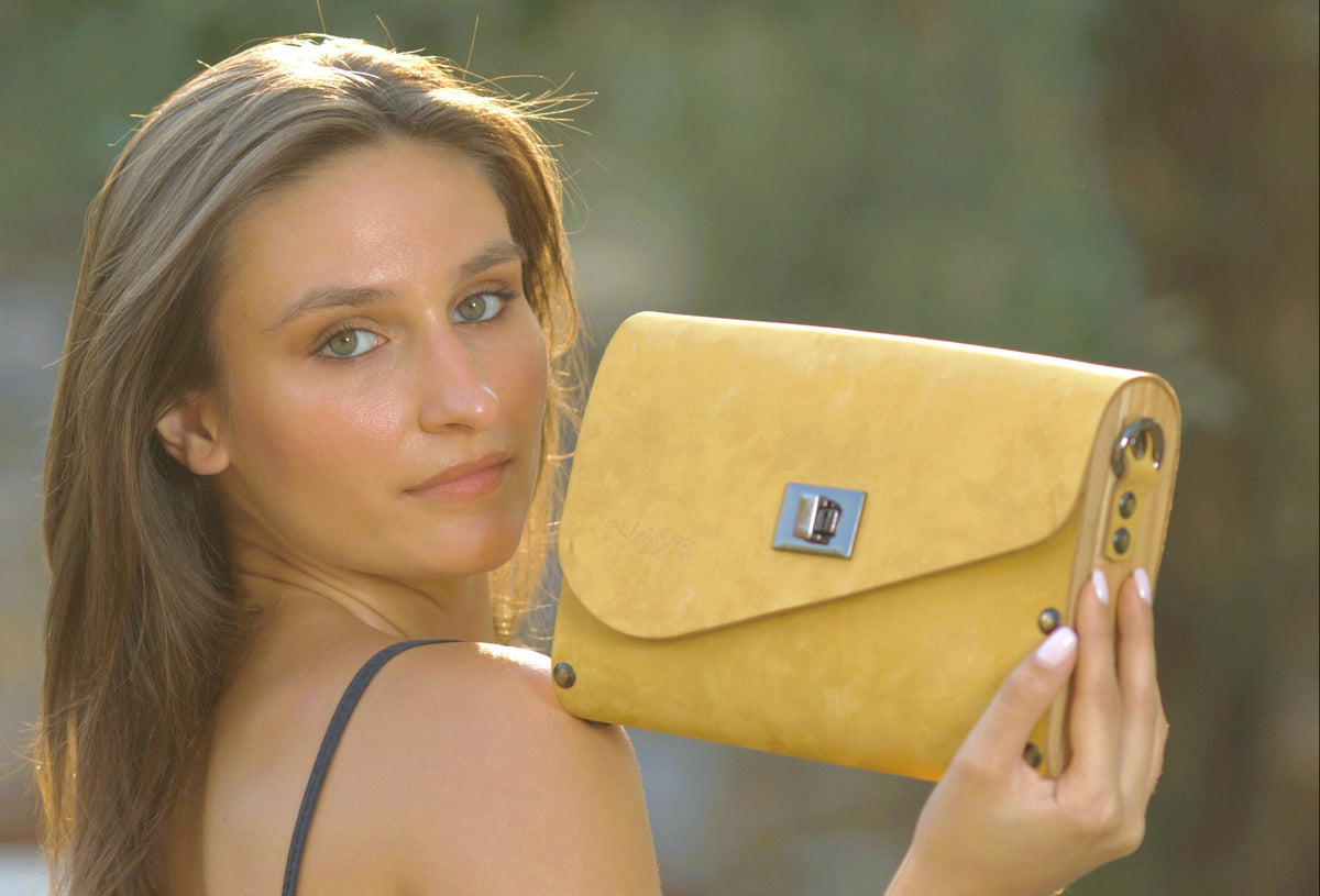 A woman holding a mustard yellow suede envelope clutch bag with a silver-tone turn-lock clasp, set against a blurred outdoor background.